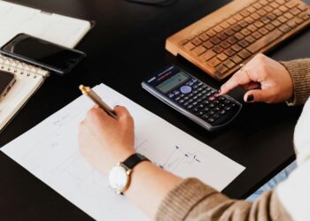 Close-up of hands working with a calculator and notebook on a desk, analyzing documents.