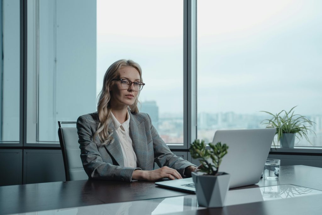 A confident businesswoman in a grey suit working on a laptop in a modern office setting.