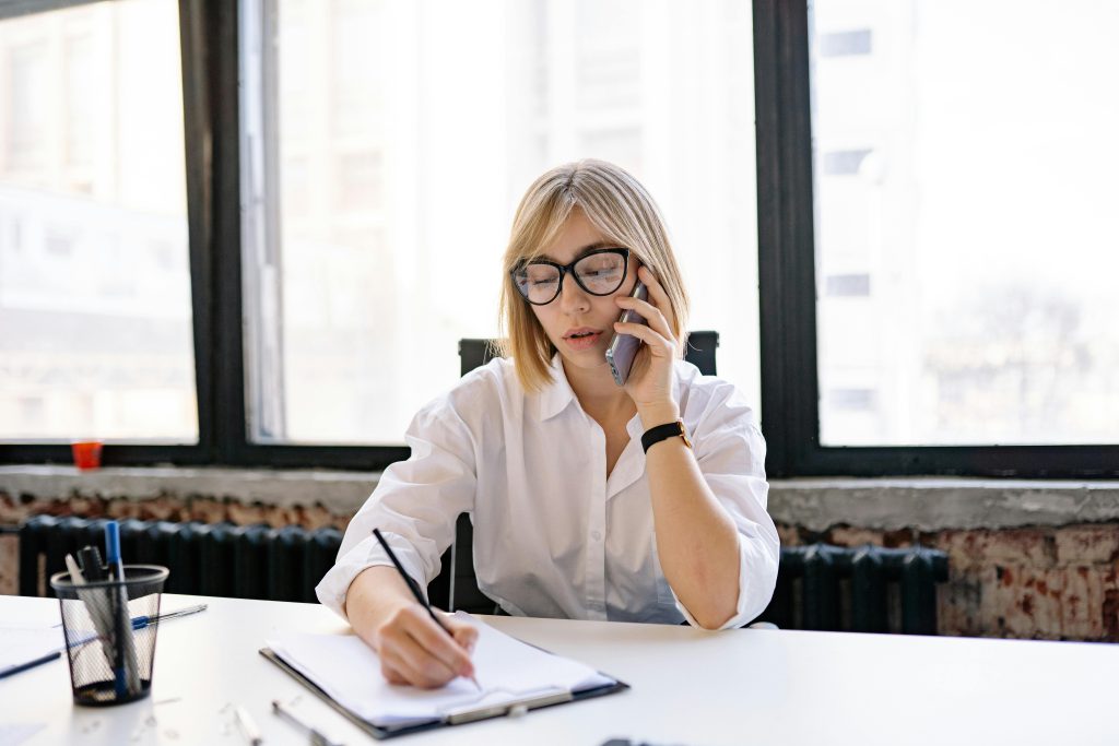 A woman in an office setting multitasking with a phone call and taking notes.
