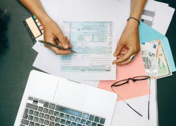 Hands writing on tax documents with laptop, glasses, and currency on desk.